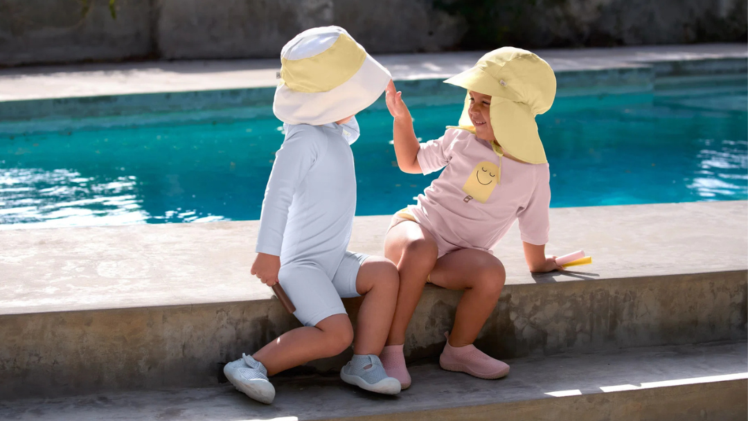 Twee kinderen zitten op de rand van een zwmebad in zwmekledij en uv werende zonnehoedjes